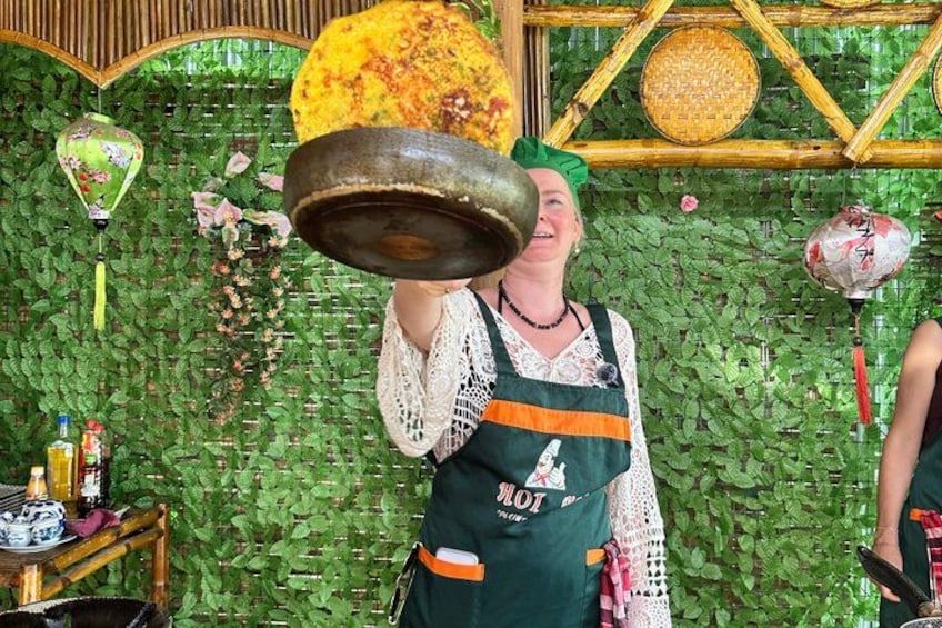 Iemand houdt een grote pot vast, draagt een groene schort met een logo, voor een groene, bladrijke wand met decoratieve details. 2