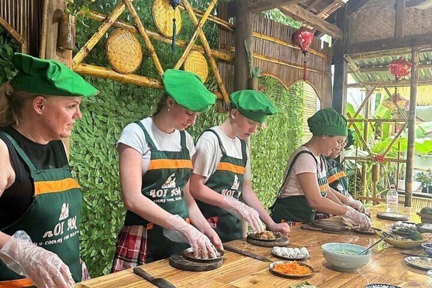 Four people wearing green hats and aprons preparing food at a wooden table with various dishes and utensils. 3
