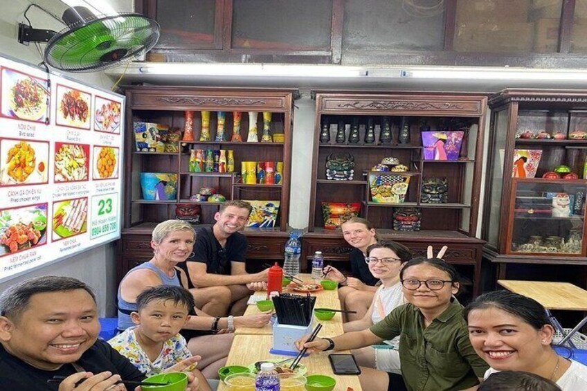 A group of people seated at a table in a restaurant with a menu on the wall and various food items on shelves. 4