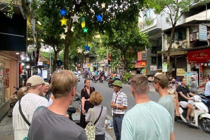 A street lined with shops and trees, with people walking and a few motorbikes. 2
