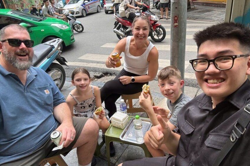 A group of people sitting at a table outdoors, eating food and holding drinks. 5