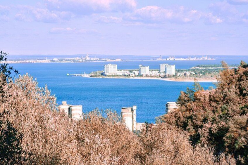 A coastal cityscape with buildings, a clear blue sea, and dry vegetation in the foreground. 5