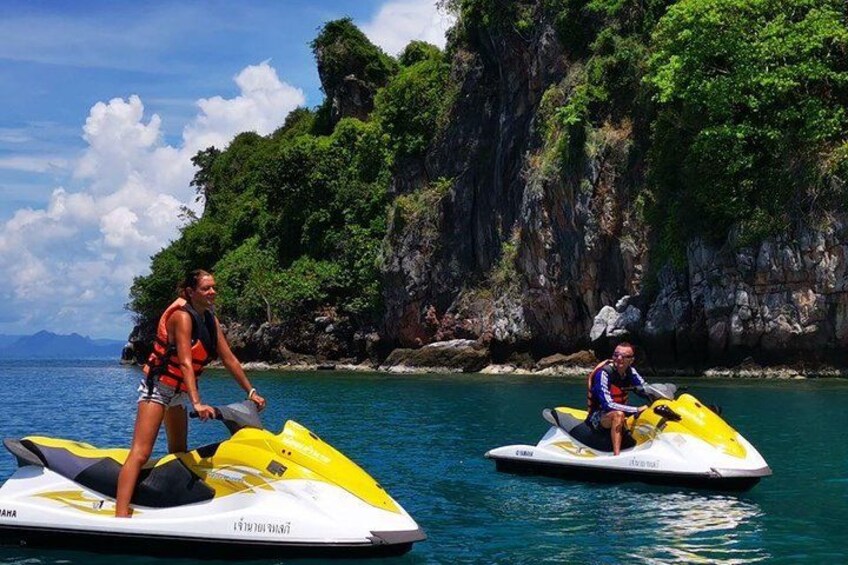 Zwei Personen auf Jetskis in einer tropischen Landschaft, mit blauem Himmel und felsigen Klippen im Hintergrund. 1