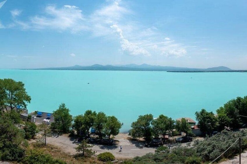 Un lago turquesa con cielo despejado, rodeado de árboles y algunos edificios. 3