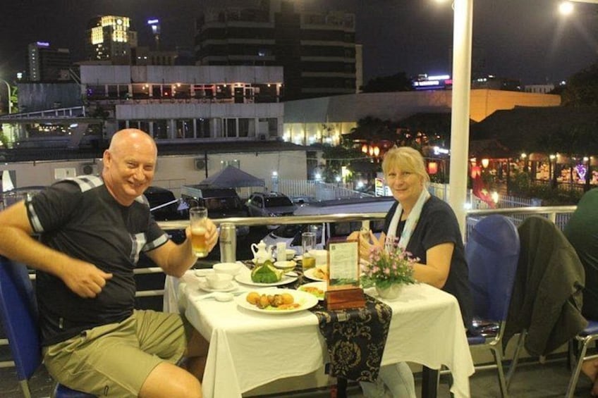 Twee personen dineren aan een buitentafel, met een stadsgezicht op de achtergrond. 5