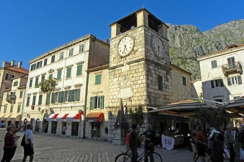 A historic stone building with a clock tower, cobblestone street, and people walking. 1