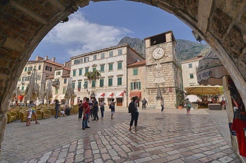 A cobblestone square with people walking, a clock tower, and a shop with a red awning. 2