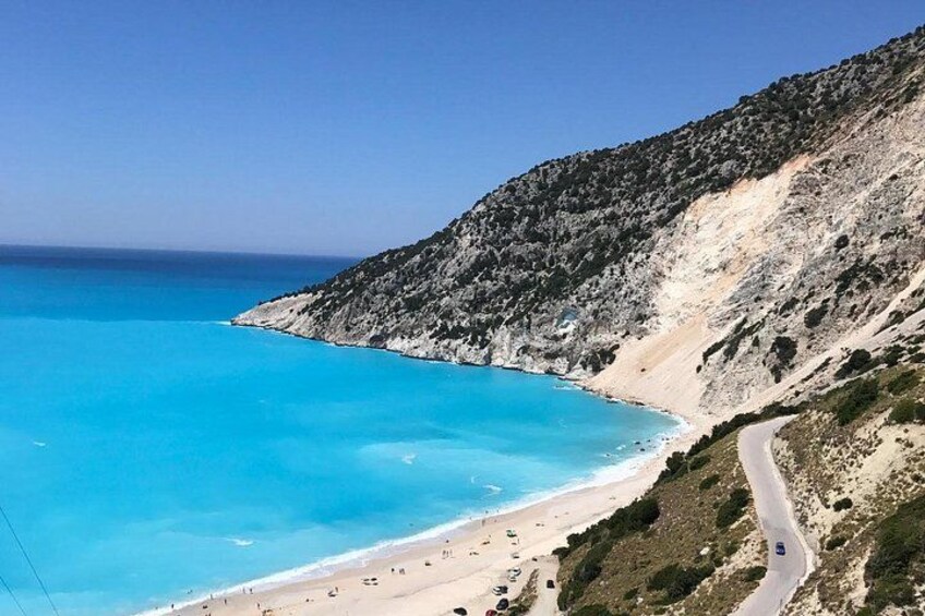 A coastal road winding along a beach with clear blue waters and rocky cliffs. 1