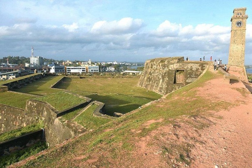 A historical fort with a clock tower, surrounded by grassy areas and a distant cityscape. 5
