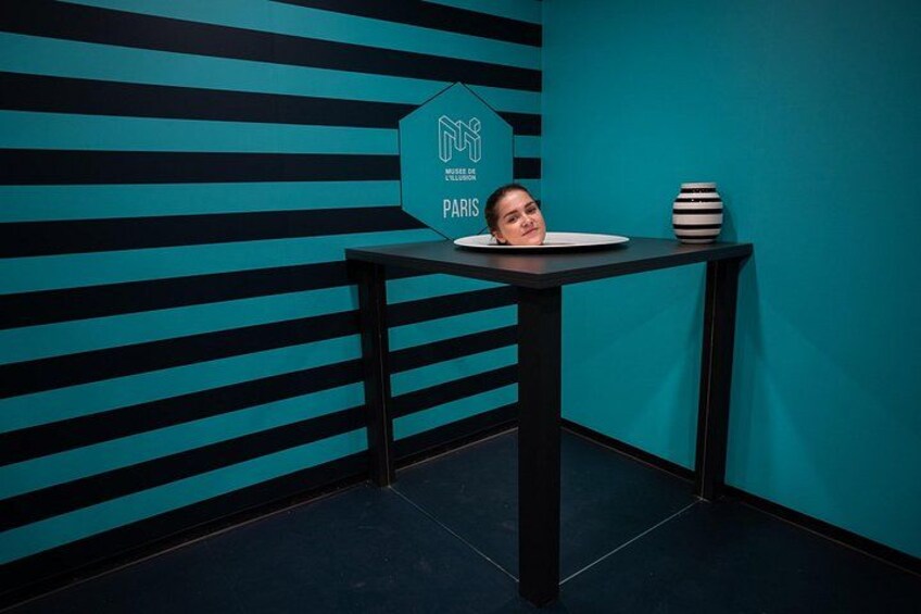 A room with a striped wall, a table with a vase, and a sign with a logo and text. 1
