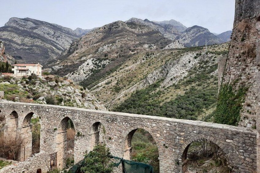 A stone bridge with arches, a mountain landscape, and a building in the distance. 1