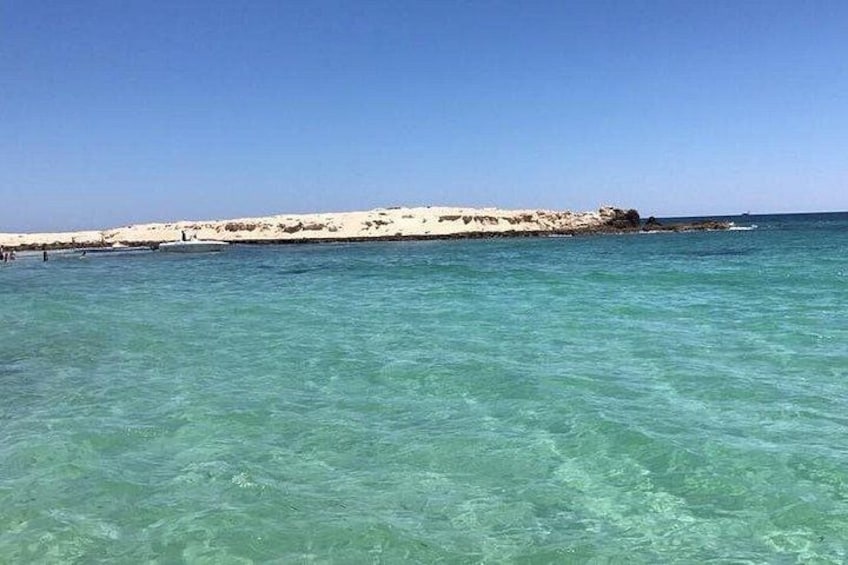 Eine Küstenlandschaft mit klarem türkisblauem Wasser, einem Sandstrand und einem felsigen Vorsprung in der Ferne. 3