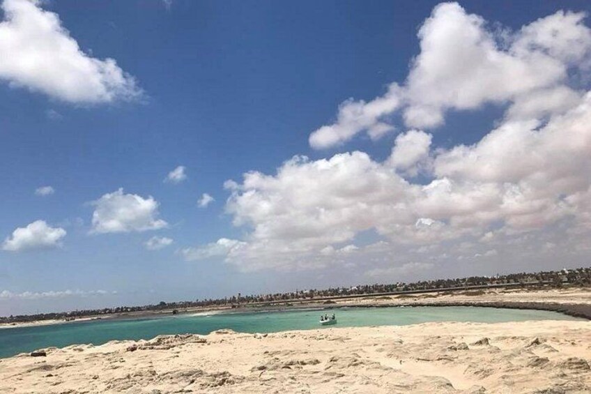 Ein Strand mit klarem türkisblauem Wasser, einem sandigen Ufer und einer fernen Küstenlinie mit Gebäuden. 2