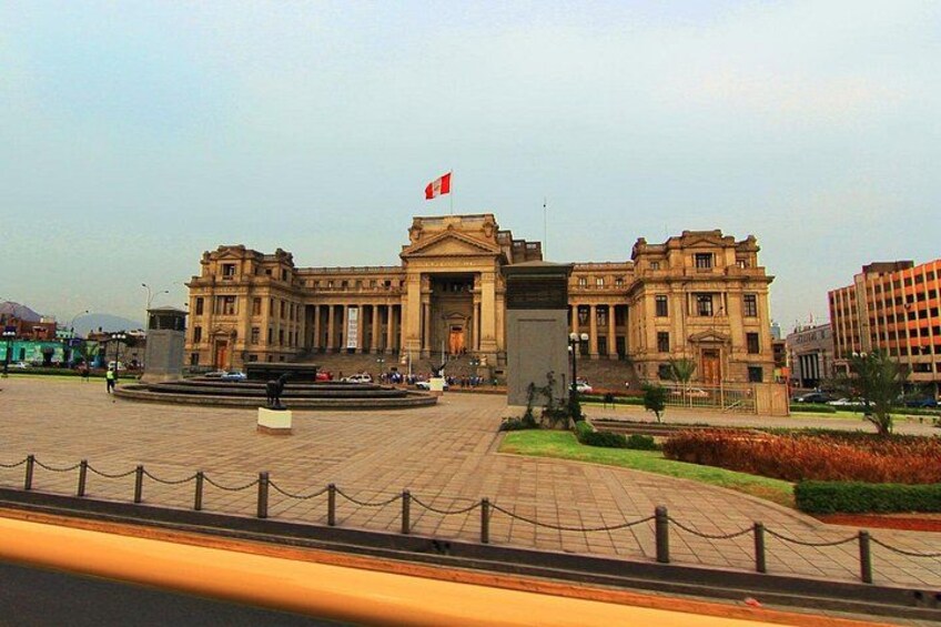 A large, historic building with a central fountain, surrounded by a paved plaza and a chain-link fence. 1