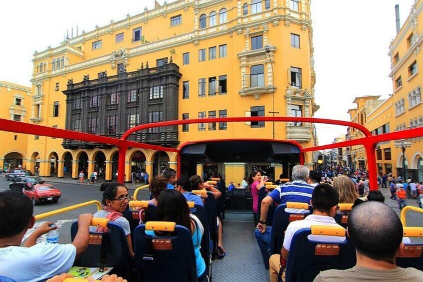 A red double-decker bus with passengers seated inside, parked in front of a yellow building with classical architecture. 4