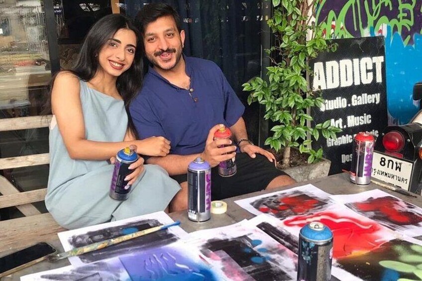 Two people sitting at a table with cans of drink, surrounded by artwork and a sign that reads 'ADDICT Studio Gallery Art Music.' 3