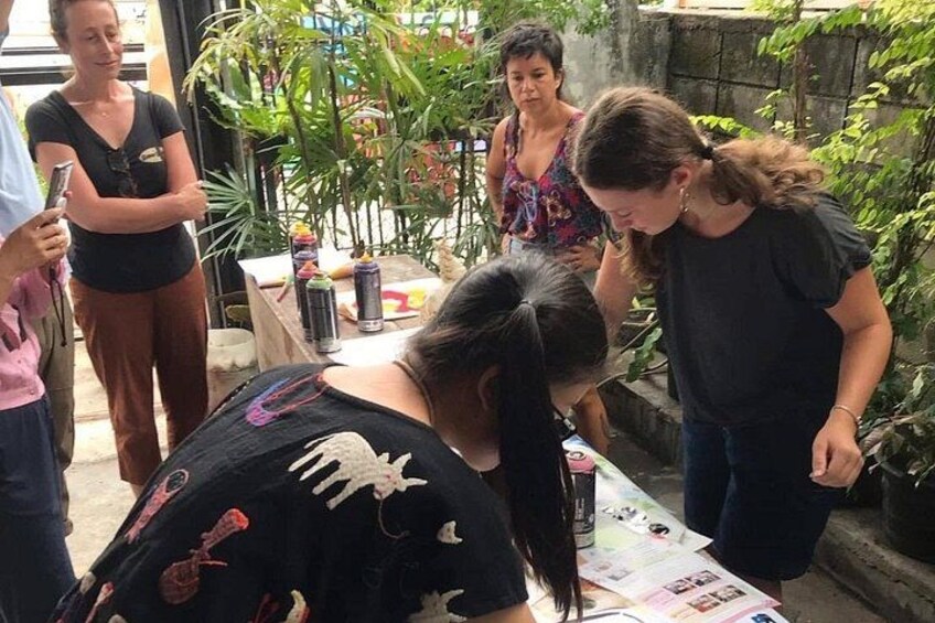 A group of people gathered around a table with various items, including a bottle and papers, in an outdoor setting with plants and a brick wall. 4