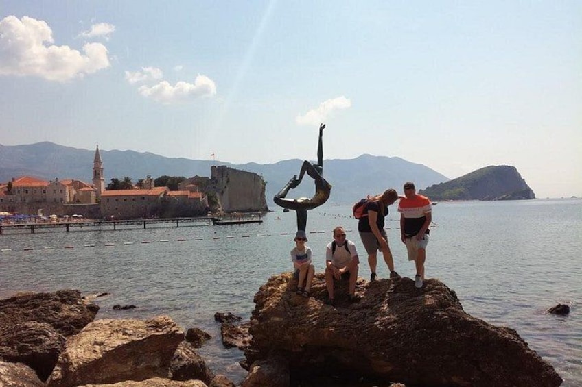 A person performing a handstand on rocks by the water, with a historic building and mountains in the background. 1