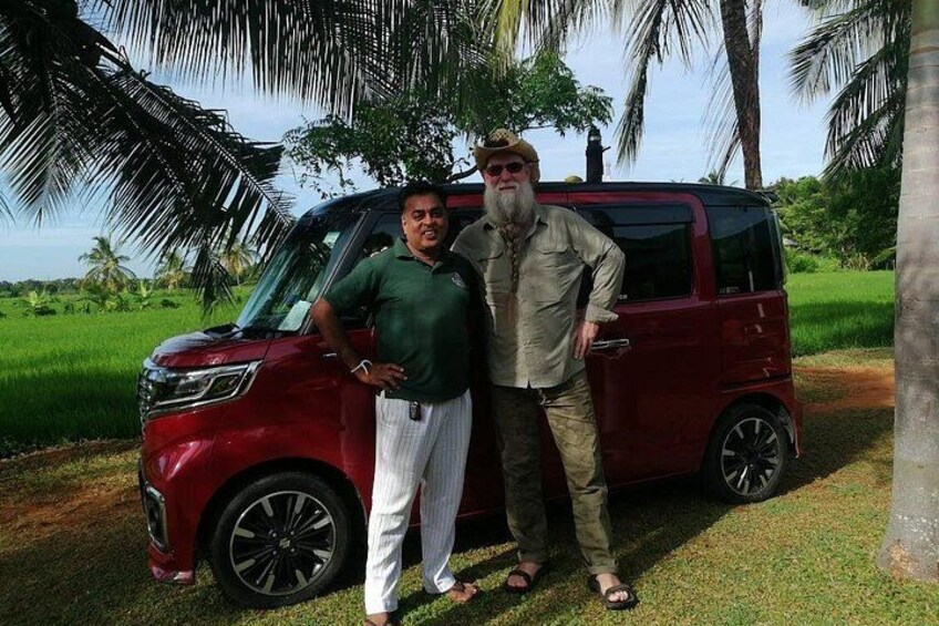 Two men standing next to a red vehicle in a grassy area with palm trees in the background. 3