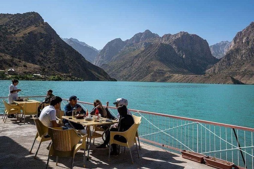 Een groep mensen dineert aan tafels met uitzicht op een turquoise meer, met bergen op de achtergrond. 2