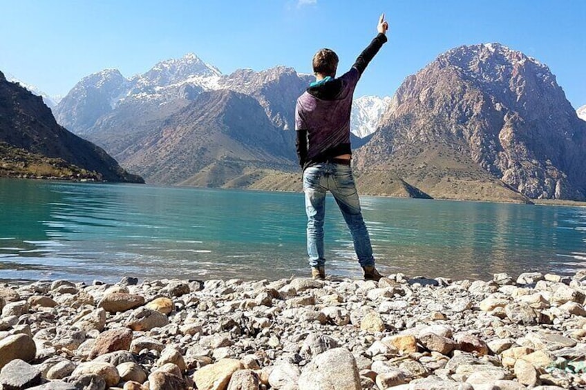 Una persona está de pie en una orilla rocosa junto a un lago, con montañas al fondo. 1