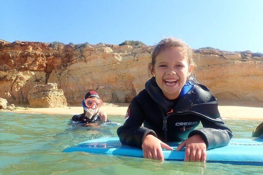 A person in a wetsuit on a blue surfboard in shallow water with rocky cliffs in the background. 3