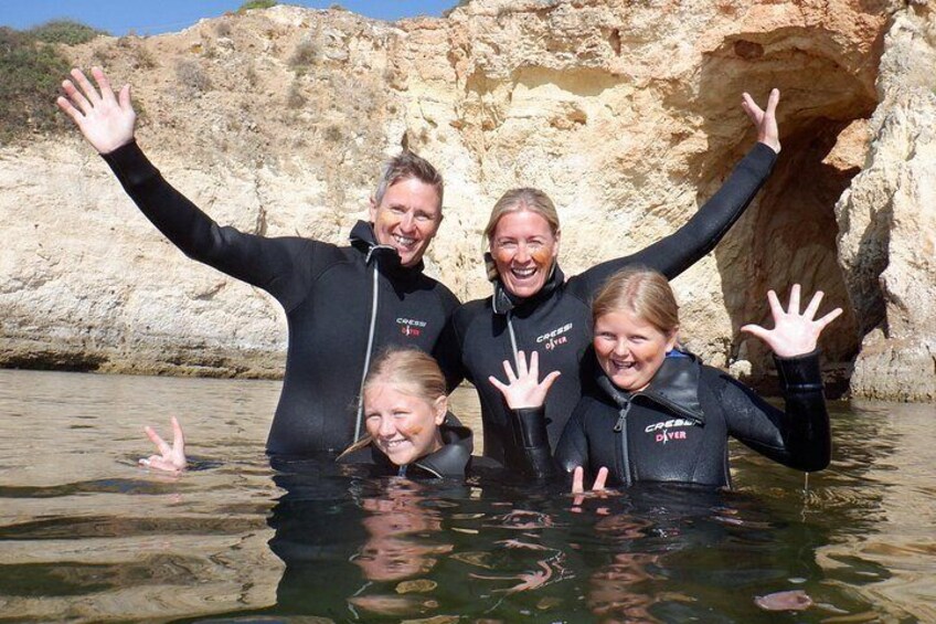Four people in wetsuits standing in shallow water with their arms raised, in front of a rocky cliff. 2