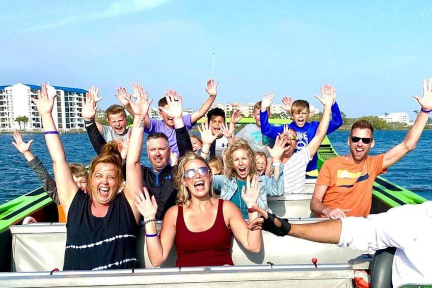 A group of people on a boat with their hands raised, facing a body of water with buildings in the background. 3