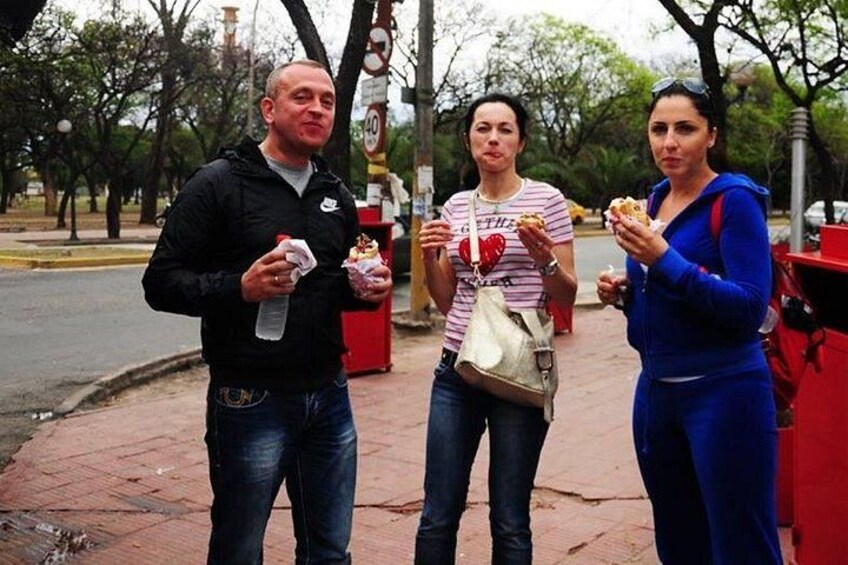 Three people eating on a sidewalk with trees and a street in the background. 2