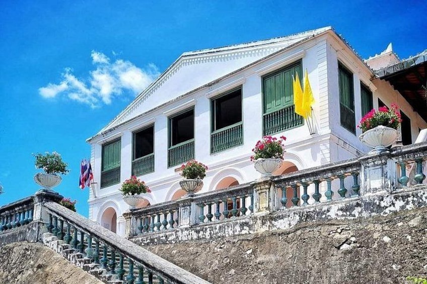A white colonial-style building with green shutters, a balcony with potted plants, and a flag. 2