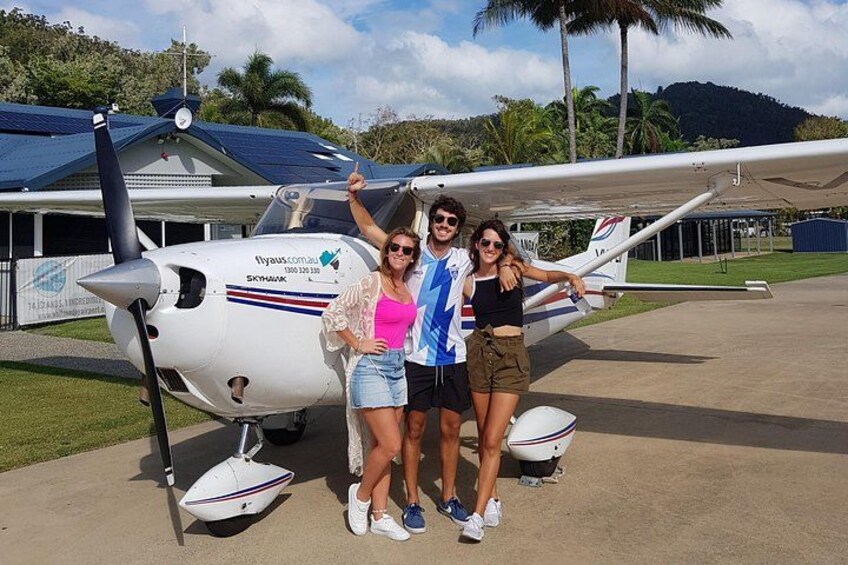 A small white airplane with a propeller parked on a tarmac, a building with a blue roof, and palm trees in the background. 2