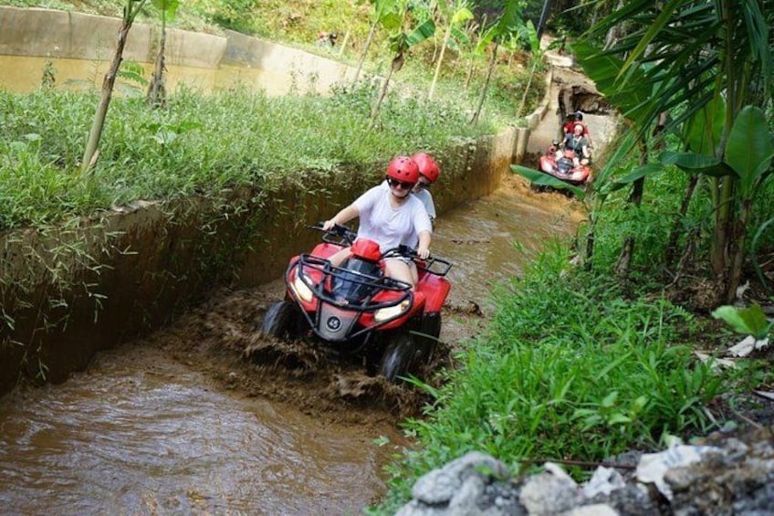 A person riding a red ATV through a muddy water channel surrounded by greenery. 5