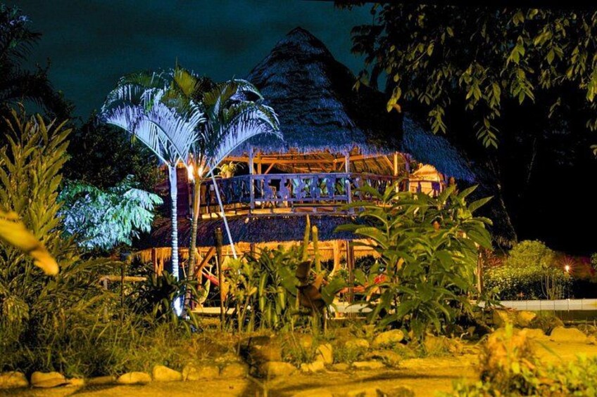 A thatched-roof building with a balcony, surrounded by tropical vegetation. 1