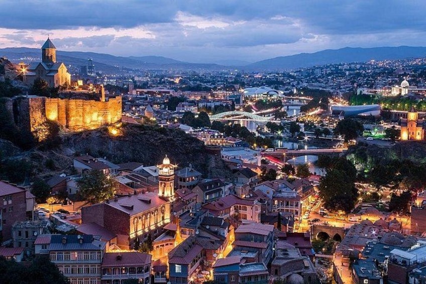 A cityscape at dusk with illuminated buildings, a prominent castle, and a bridge. 5