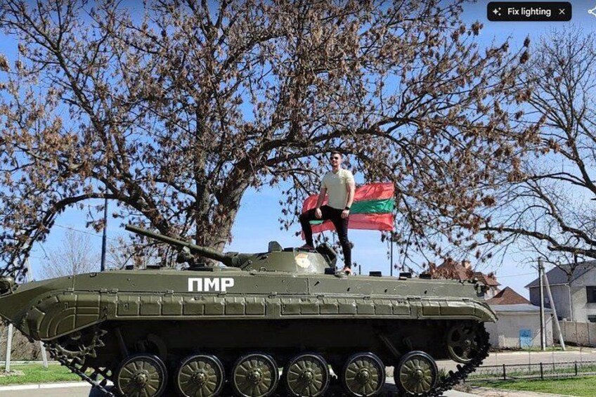 Un tanque militar con una persona de pie encima, una bandera y árboles al fondo. 1