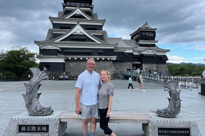 A historic castle with a traditional Japanese architectural style, a stone bench, and two people standing in front. 1
