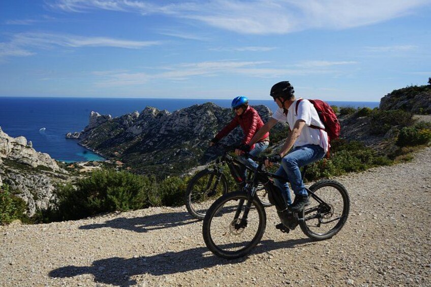 Two people riding bicycles on a gravel path with a mountainous landscape and the sea in the background. 2