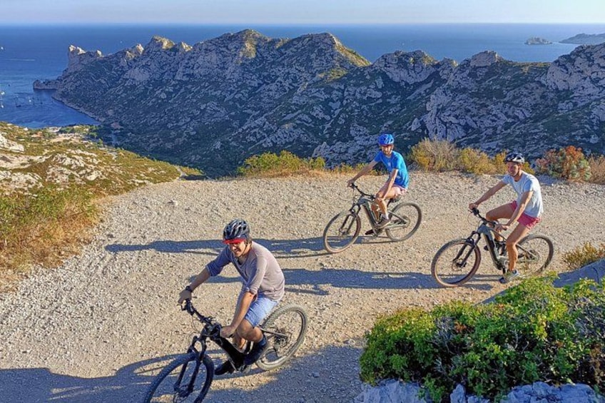 Three cyclists on a gravel path with a mountainous landscape and the sea in the background. 1
