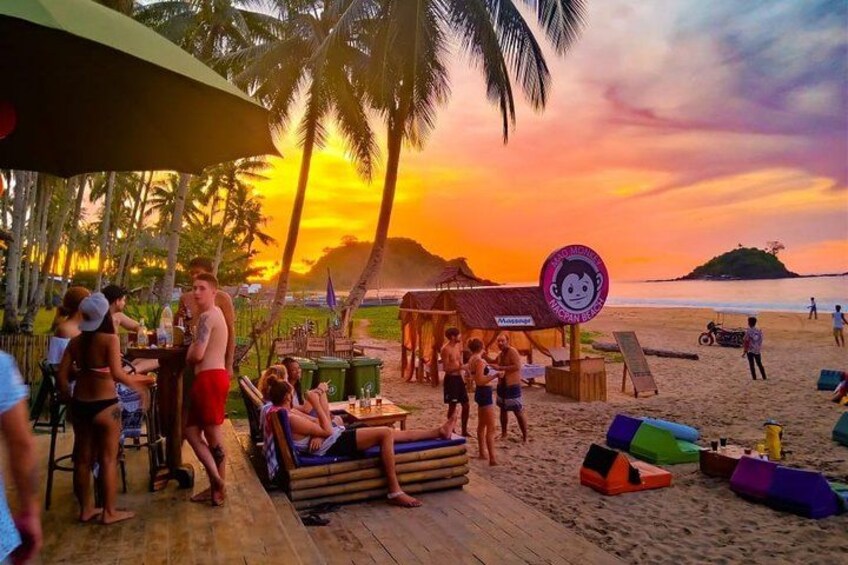 A beach scene with people relaxing, a wooden deck, lounge chairs, and palm trees. 5