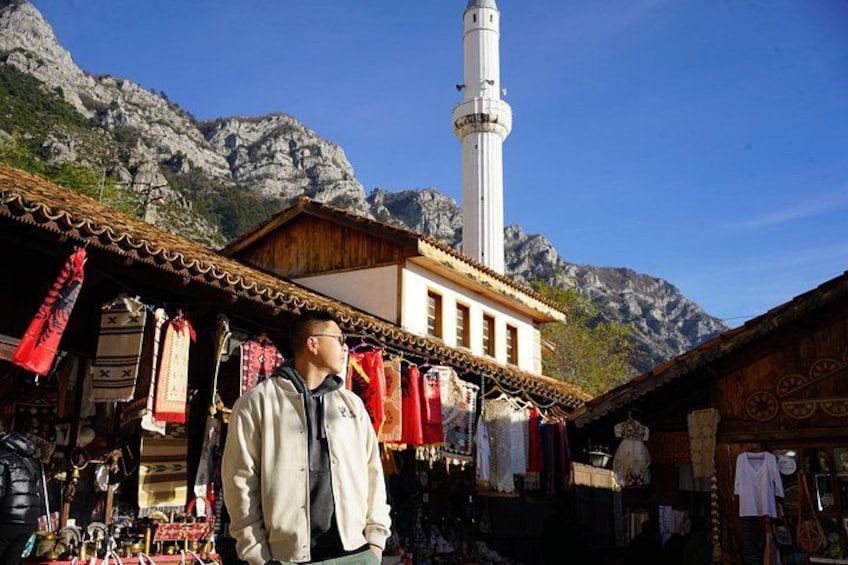 Eine Person steht vor einem traditionellen Gebäude mit einem Minarett, umgeben von gebirgiger Landschaft. 1
