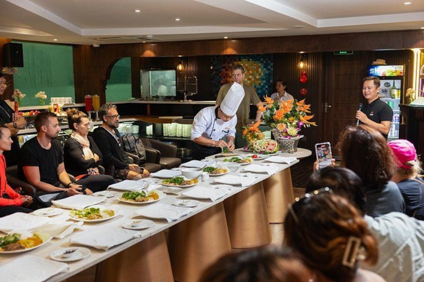 Un chef cocina frente a un público sentado en una larga mesa. 4
