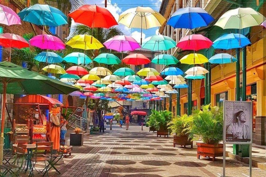 A street with colorful umbrellas hanging overhead, a market stall, and a sign with a woman's portrait. 1