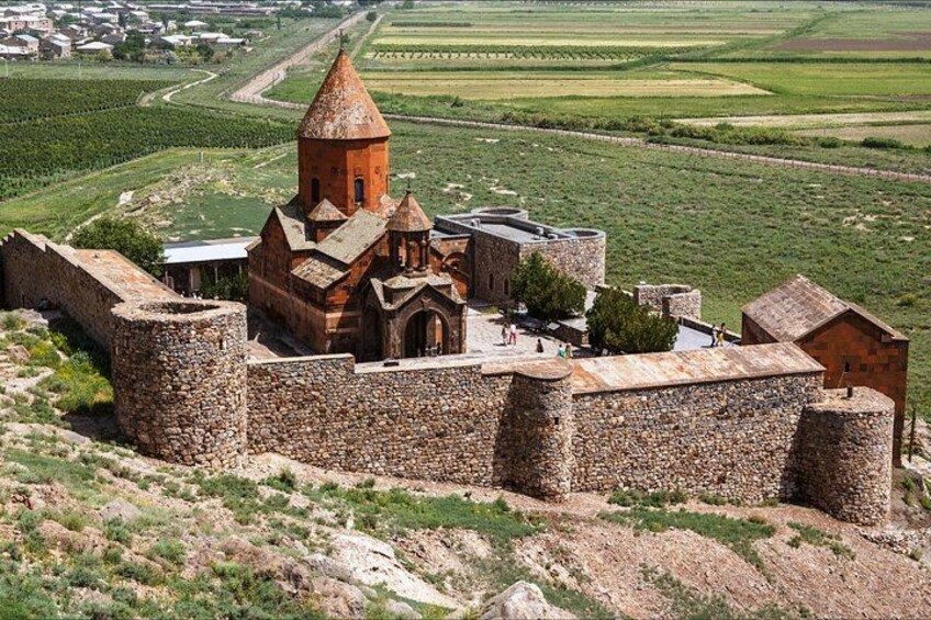 A historic stone fortress with a prominent tower, surrounded by lush green fields and a winding road in the background. 4