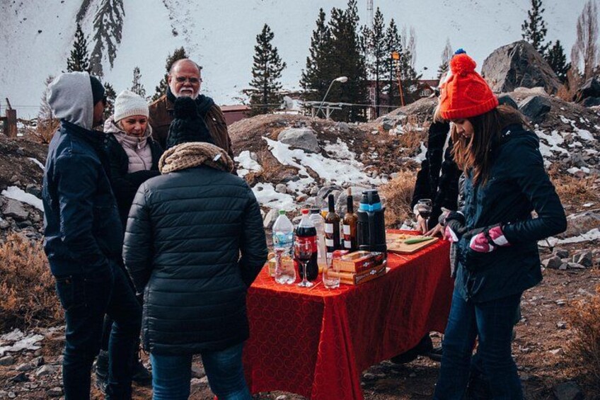 A group of people around a table with bottles and glasses, outdoors with snow and trees in the background. 1