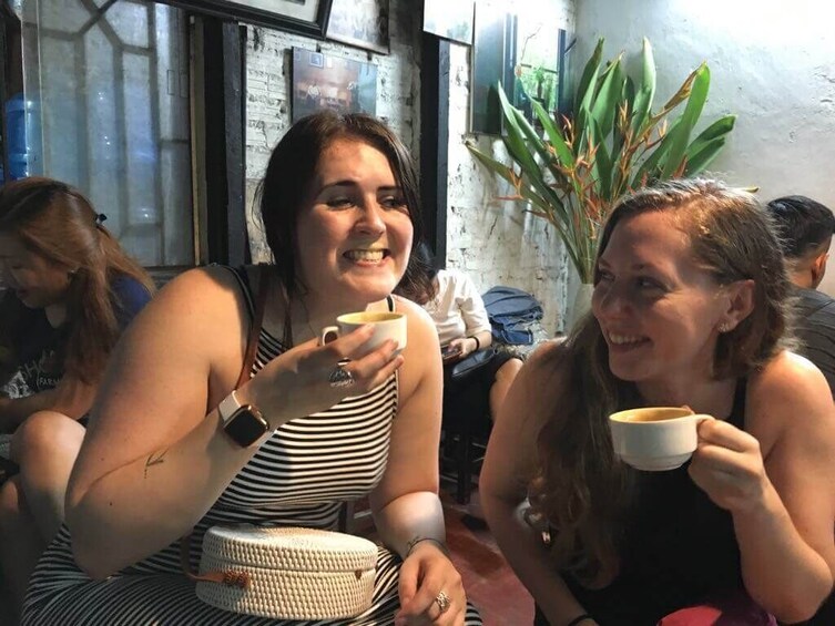 Due donne sedute a un tavolo, ognuna con una tazza in mano, in un caffè con una pianta sullo sfondo. 4