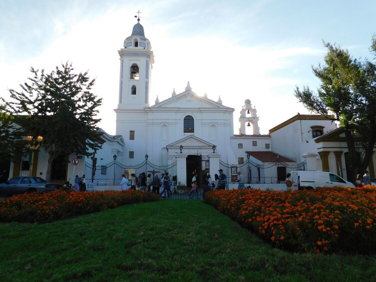 A white church with a bell tower and a clock. There are orange flowers in the foreground and people walking around. 1