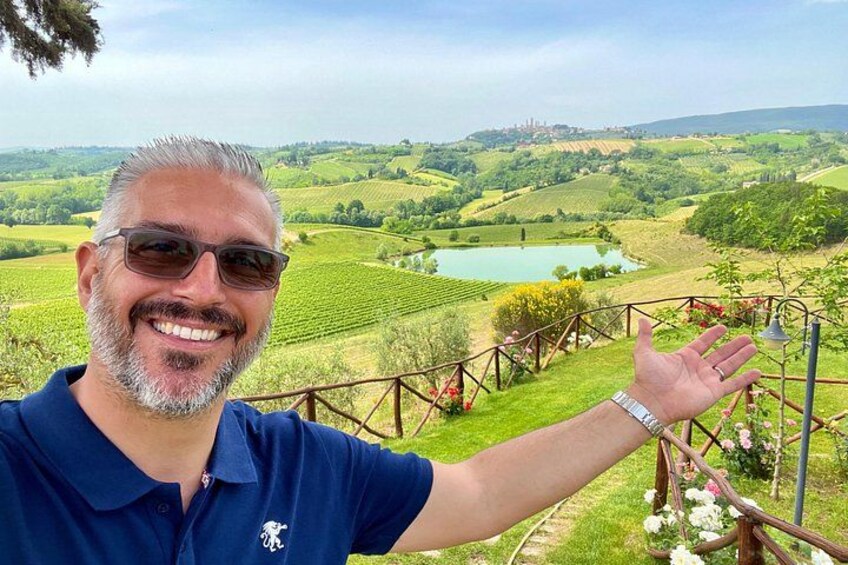A person standing with a hand raised, overlooking a vineyard with rows of grapevines and a body of water in the distance. 5