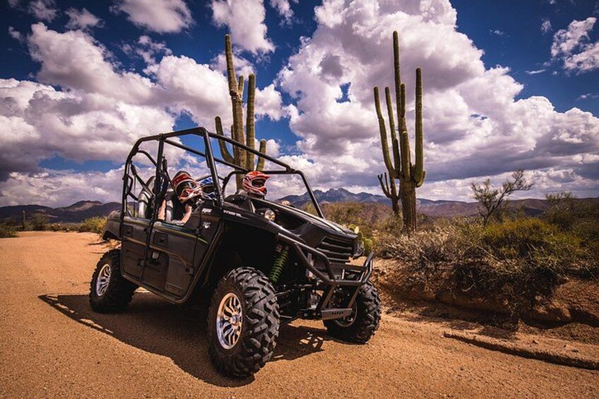 A black off-road vehicle with two people wearing helmets, parked on a dirt road with cacti and mountains in the background. 2