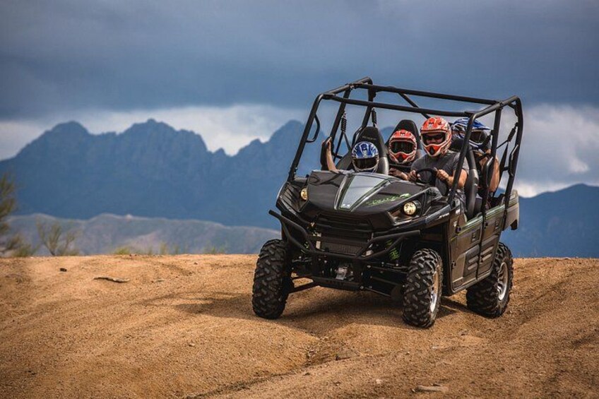 A four-wheeled off-road vehicle with two passengers wearing helmets, driving on a dirt road with mountains in the background. 3