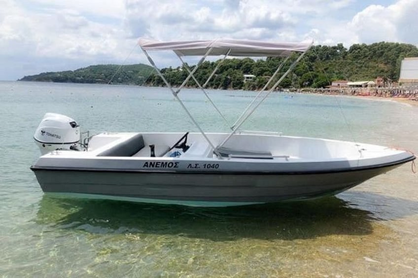A boat named 'ANEMOZE' with a canopy, on a clear water body with a beach and greenery in the background. 2